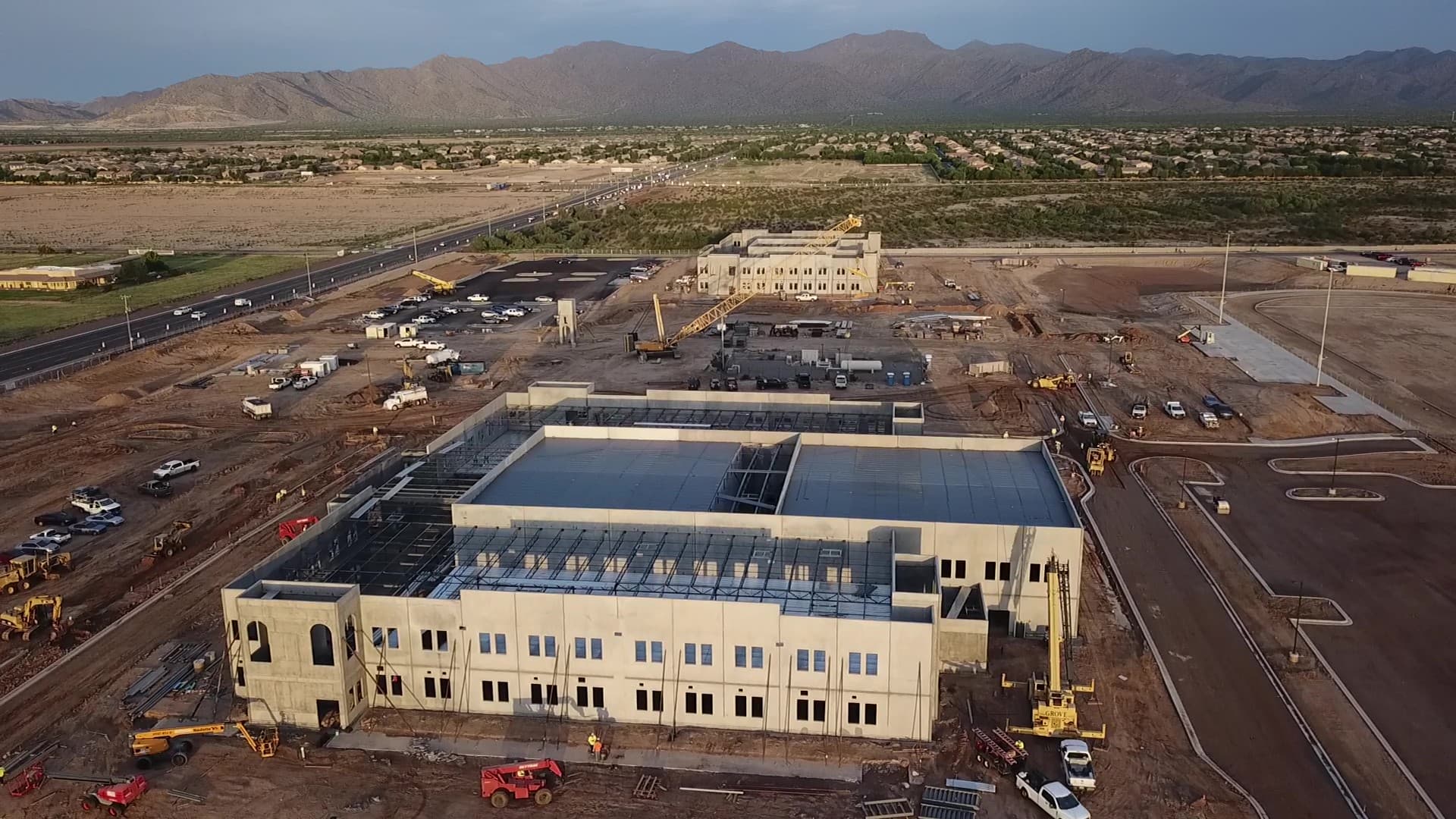 Aerial view of a large construction site with concrete buildings, cranes, and distant desert mountains.