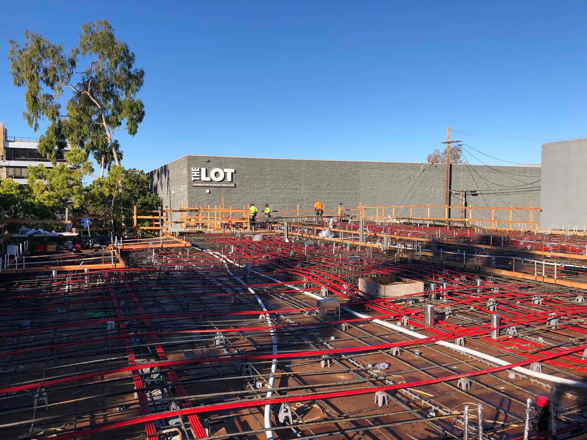 Construction workers install rebar and red conduits on a platform near The Lot building.