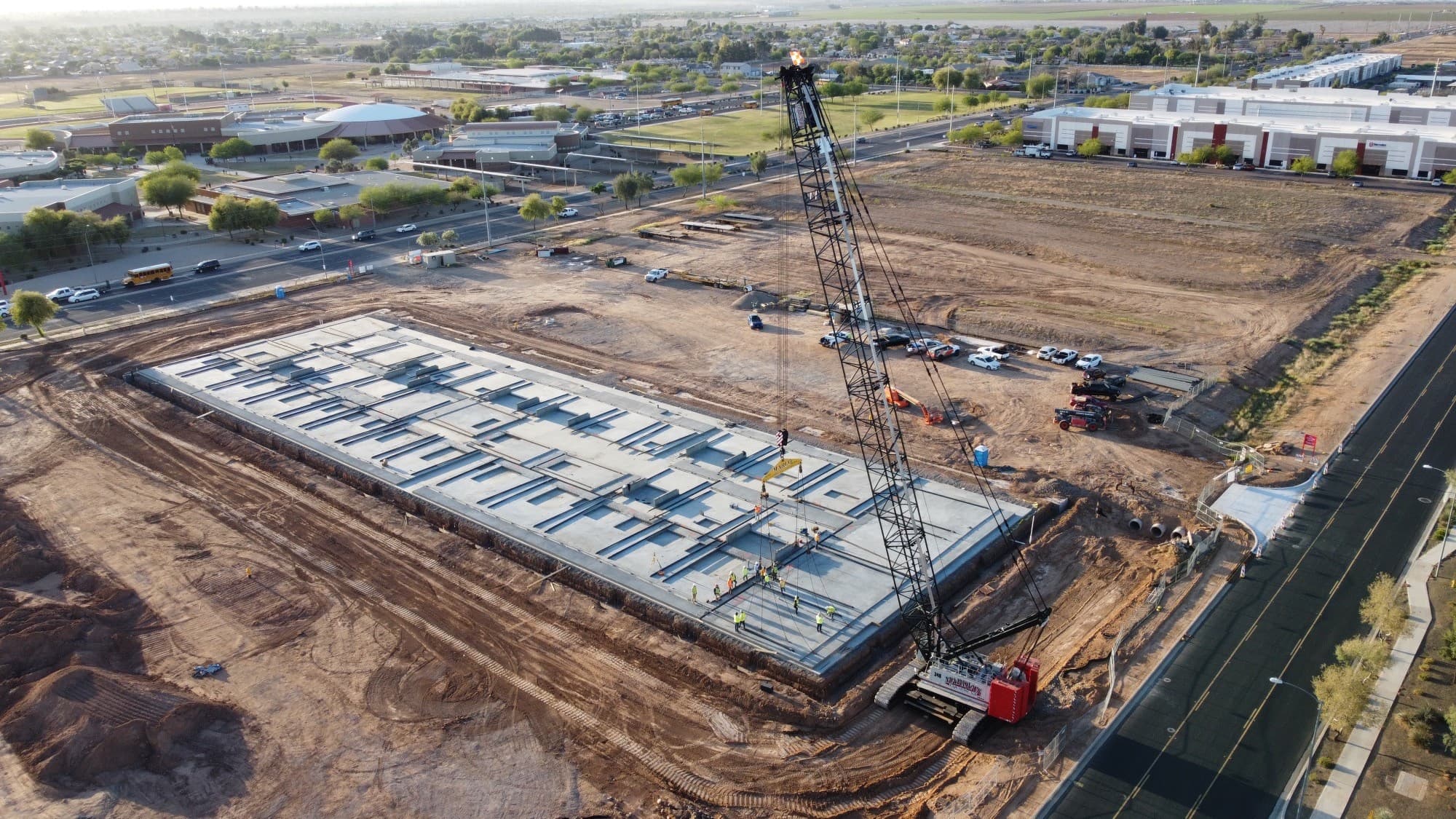 Tall black crane and workers on a large concrete foundation at a suburban construction site.